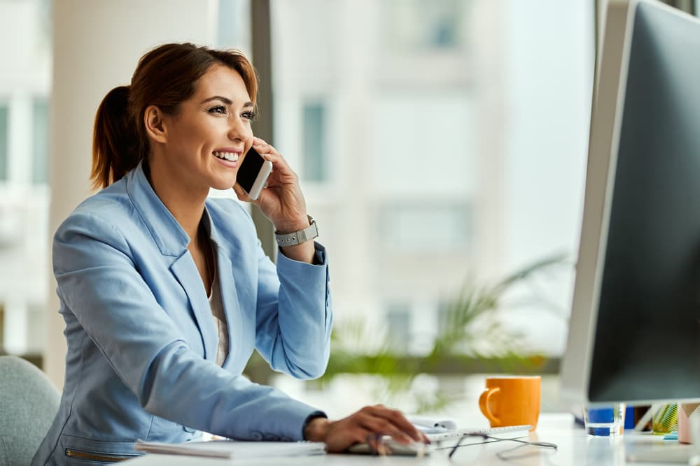 A photograph of a young adult female working on a computer while having a conversation on her cell phone